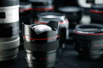 Imagem mostrando vários tipos de lentes para câmeras fotográficas DSLR na posição vertical sobre uma mesa com fundo escuro. 