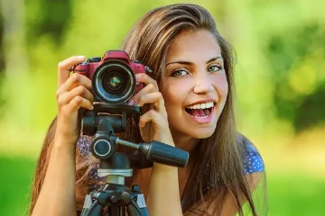 Mulher sorrindo ao segurar uma câmera sobre um tripé em ambiente natural externo.